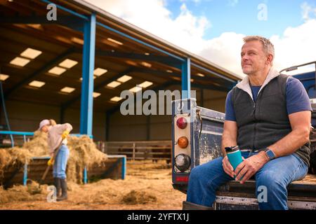 Reifer Männlicher Landarbeiter Sitzt Auf Der Heckklappe Eines Geländefahrzeugs Und Hält Heißgetränk Im Cup Stockfoto