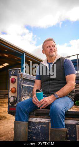 Reifer Männlicher Landarbeiter Sitzt Auf Der Heckklappe Eines Geländefahrzeugs Und Hält Heißgetränk Im Cup Stockfoto