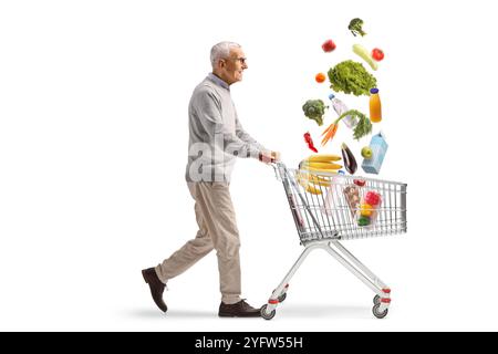 Profilaufnahme eines älteren Mannes, der einen Einkaufswagen mit herabfallenden Lebensmittelprodukten auf weißem Hintergrund läuft und schiebt Stockfoto