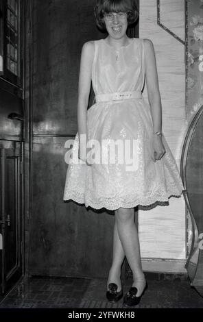 1950er Jahre, historisch, ein großes Lächeln ..inside ein Flur, eine junge Frau in einem glänzenden, ärmellosen Partyrock, die für ein Foto steht, ein Kreuz, das an einer Kette am Hals hängt, England, Großbritannien. Stockfoto