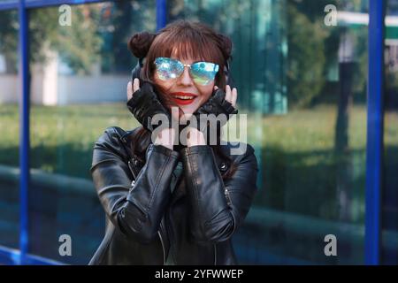 Ein wunderschönes Mädchen in einer schwarzen Lederjacke und Handschuhen, trägt eine blaue Sonnenbrille und hält vor den großen blauen Fenstern Kopfhörer auf dem Kopf. Stockfoto