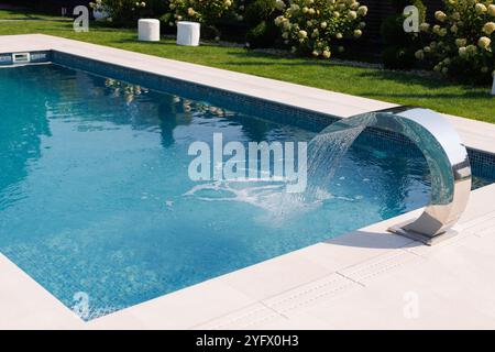 Ein moderner Swimmingpool mit einem schlanken, geschwungenen Wasser gießt Wasser in den Pool. Der Pool hat eine klare blaue Wasseroberfläche und ist umgeben von gr Stockfoto