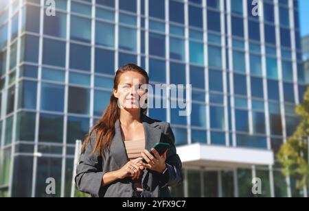 Eine Geschäftsfrau in einem Business-Outfit hält ein Smartphone, während sie auf einer Stadtstraße läuft. Stockfoto