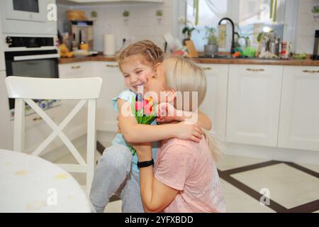 Ein freudiger Moment zwischen einer Mutter und ihrer Tochter, während sie einander umarmen und lebendige Blumen in den Händen halten. Vor dem Hintergrund einer hellen, weißen Küche strahlt ihr Lächeln Liebe und Glück aus. Stockfoto