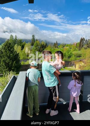 Eine Gruppe von Kindern genießt einen sonnigen Tag auf einem Holzbalkon mit Blick auf üppiges Grün und lebhaften Himmel. Ein friedlicher und fröhlicher Familienmoment im Freien Stockfoto