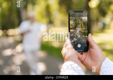 Nahaufnahme eines bärtigen älteren Mannes, der sich glücklich im sonnigen Park posiert, während die liebende Frau Fotografin im sonnigen Sommerpark Momente mit dem Smartphone festnimmt. Stockfoto