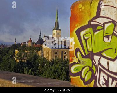 Blick auf die Altstadt von Tallinn mit der markanten St.-Olev-Kirche und historischen Gebäuden, grüne Baumkronen im Vordergrund, kontrastiert von einem bunten Graffiti auf einer Betonwand rechts im Bild, lebendige Street-Art trifft auf mittelalterliche Architektur, symbolisiert den Mix aus Tradition und Moderne in Tallinn, Abendlicht verstärkt die Farben und verleiht der Szene eine warme Atmosphäre, Symbolbild *** Blick auf die Altstadt von Tallinn mit der markanten St. Olev Kirche und historischen Bauten, grüne Baumkronen im Vordergrund, im Kontrast dazu steht ein farbenfrohes Graffiti an einer Betonwand an der Stockfoto