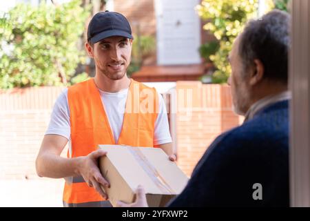 Junger Mann, der als Liefermann arbeitet und eine Kiste zu Hause an Kunden liefert. Freundlicher Mitarbeiter, der den Kunden lächelt. Stockfoto