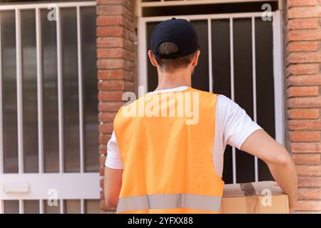 Ein junger und attraktiver Mann arbeitet als Paketlieferant. Städtischer Hintergrund mit Fahrer, der vor der Tür des Kunden wartet Stockfoto