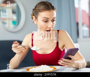 Unzufriedene Frau, die Reis isst und gleichzeitig eine Nachricht am Telefon tippt, während sie am Tisch im Zimmer sitzt Stockfoto