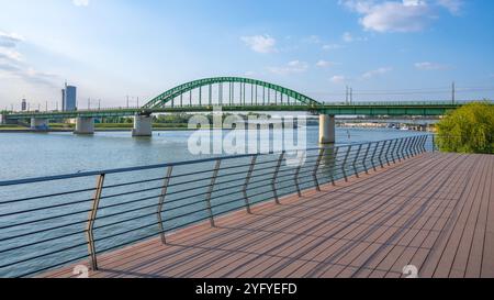 Die alte Save-Brücke überspannt die Save in Belgrad mit ihren grünen Bögen. Eine hölzerne Promenade verläuft entlang des Flusses, die die ruhige Atmosphäre am Wasser während des Sonnenuntergangs unterstreicht. Stockfoto