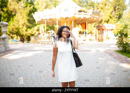 Nachdenkliche Frau mit Vitiligo-Hautstörung, die Handtasche beim Stehen im Vergnügungspark trägt. Eine junge Frau in weißem Kleid schaut während des sonnigen Tages weg. Stockfoto