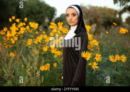 Nachdenkliche ernste Nonne, die während des Tages auf dem Feld steht. Die junge Frau in religiöser Kleidung ist im Freien von frischen gelben Blumen umgeben. Stockfoto