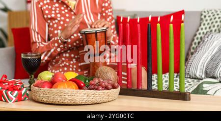 Brennende Kerzen mit Essen und Geschenke für Kwanzaa auf dem Tisch und eine Frau, die zu Hause tam-tam-Trommel spielt Stockfoto