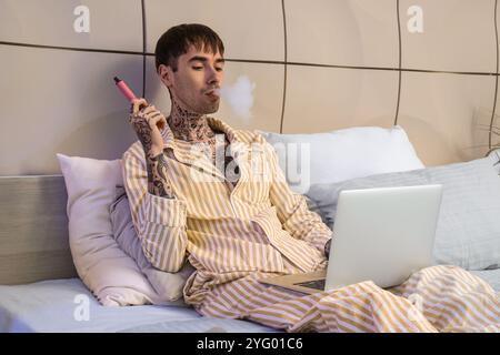 Junger Mann im Schlafanzug mit elektronischer Einwegzigarette mit Laptop auf dem Bett zu Hause Stockfoto