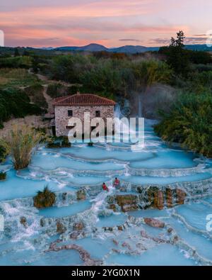 Besucher entspannen sich im beruhigenden, türkisfarbenen Wasser der Saturnia Thermalbäder, während die Sonne hinter der üppigen toskanischen Landschaft untergeht und eine magische Atmosphäre schafft Stockfoto