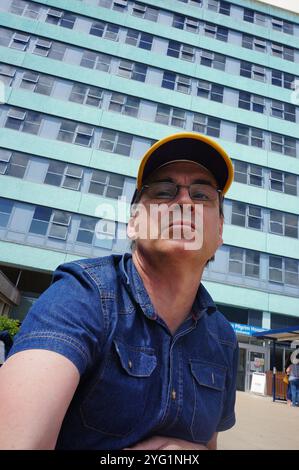 Ein reifer Mann mit Baseballmütze und Brille, der vor einem mehrstöckigen Gebäude steht. Stockfoto