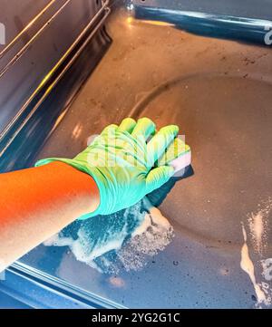 Weibliche Hand im grünen Gummihandschuh, der den schwarzen Ofen mit rosa Schwamm reinigt. Hochwertige Fotos Stockfoto