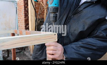 Ein Mann in schmutziger Kleidung mit einem Schraubenzieher schraubt selbstschneidende Schrauben in eine Holzkonstruktion, die sie mit einer Hand auf der Straße hält Stockfoto