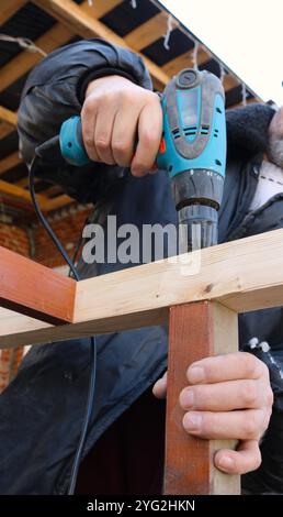 Der Arbeiter verwendet einen Schraubendreher, um eine selbstschneidende Schraube in eine Holzkonstruktion zu bohren oder zu Schrauben, während er sie mit einer Hand hält Stockfoto