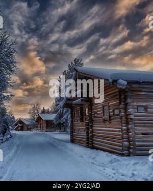 Eine Reihe von Blockhütten befindet sich auf einer schneebedeckten Straße. Der Himmel ist bewölkt und die Sonne untergeht, was eine stimmungsvolle Atmosphäre schafft Stockfoto