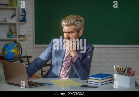 Porträt eines selbstbewussten männlichen Lehrers im Klassenzimmer mit Laptop in der Klasse sitzt am Tisch die Tafel im Klassenzimmer. Lehrer nachdenklich im Klassenzimmer Stockfoto