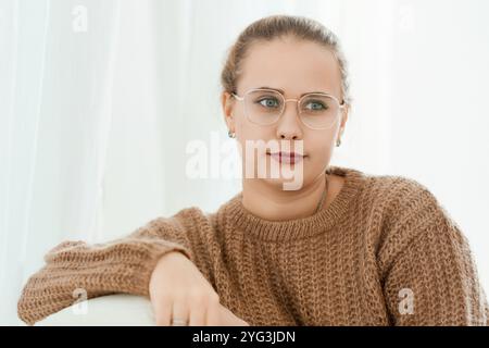 Eine nachdenkliche Frau in Brille und braunem Pullover sitzt auf einem Sofa auf hellem Hintergrund Stockfoto