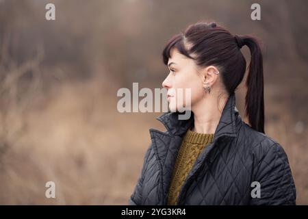 Eine Frau mit Pferdeschwanz und Ohrringen steht im Park. Sie trägt eine schwarze Jacke und einen Strickpullover Stockfoto