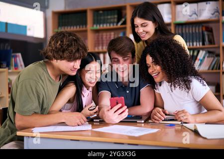 Verschiedene Gruppen von Schülern, die gemeinsam in der Bibliothek auf das Handy lächeln. Stockfoto