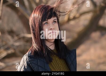 Frau mit langen Haaren und schwarzer Jacke draußen. Sie hat einen ernsthaften Gesichtsausdruck, der ihr die Haare aus dem Gesicht bläst Stockfoto