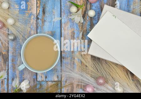 Weißer Kaffee zwischen Boho-Dekorationen, Weihnachtskugeln, leerem Blatt Papier und Umschlag zum Füllen mit Inhalt auf blauen Holztafeln. Stockfoto