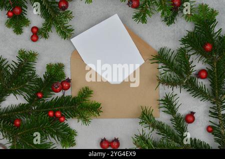 Leere Karte und Umschlag umgeben von Weihnachtsdekorationen, Weihnachtsbaumzweigen und roten Früchten, Blick von oben. Stockfoto