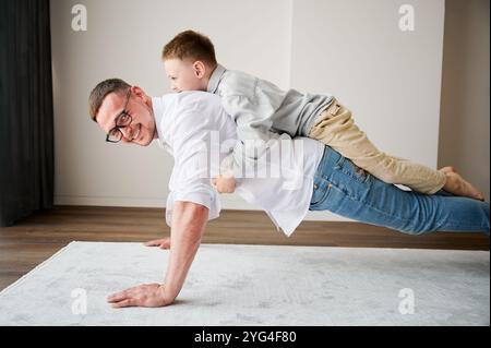 Ein energiegeladener Vater und sein kleiner Sohn trainieren zu Hause. Der Junge kletterte Dad auf den Rücken, während er Plank machte. Junge Familie, die zu Hause Aktivitätsspiele genießt. Stockfoto