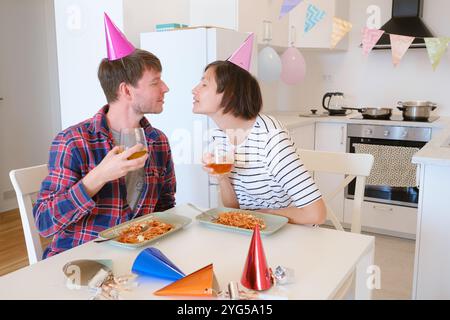 Das junge Paar feiert zu Hause gemütlich Geburtstag und genießt Kuchen und Lachen in warmer Atmosphäre. Die Familie isst Pasta und trinkt Wein, um zu feiern, zu essen und zu jubeln Stockfoto