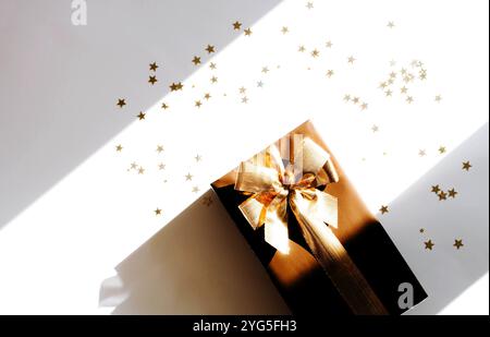 Weihnachtsgeschenkbox mit Blick von oben, mit leuchtendem goldenem Band, umgeben von goldenem sternförmigem Konfetti auf weißem Hintergrund mit Schatten. Flache Lagen. Stockfoto