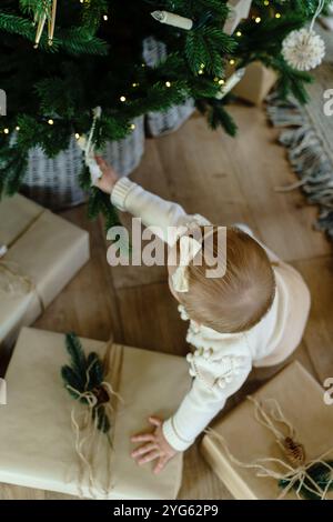 Ein kleines Mädchen sitzt am Weihnachtsbaum und greift nach einem Weihnachtsbaum Stockfoto