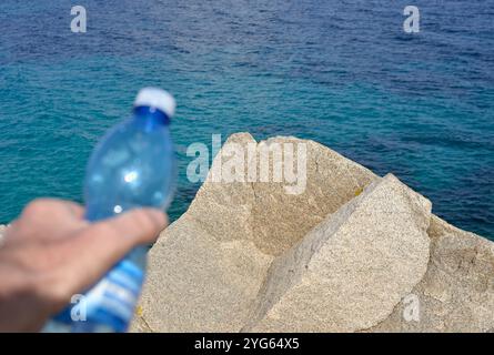 Hand hält Plastikwasserflasche vor dem Meer an einer felsigen Küste Stockfoto