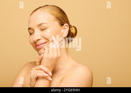 Eine strahlende Frau lächelt sanft, während sie Creme auf ihr Gesicht aufträgt und ihre Schönheit feiert. Stockfoto
