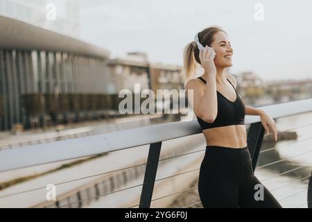 Während die Sonne über dem Wasser untergeht, lehnt sich eine junge Frau an ein Geländer und genießt ihre Lieblingsmusik über Kopfhörer, um Ruhe und Frieden zu genießen Stockfoto