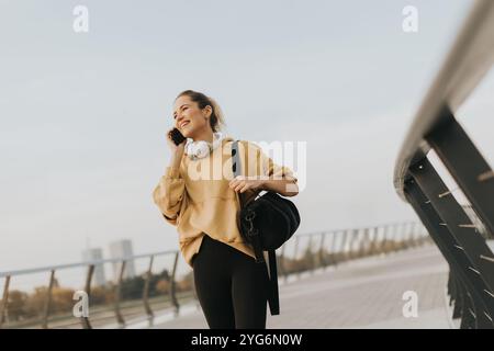Die fröhliche junge Frau schlendert entlang eines Flussufers, führt ein lebhaftes Telefongespräch, während sie ein hellgelbes Sweatshirt trägt und ein B trägt Stockfoto