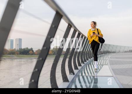Eine junge Frau in einer leuchtend gelben Jacke lehnt sich an ein modernes Geländer, während sie sich auf ihr Handy konzentriert und einen ruhigen Spaziergang am Fluss auf einem Pett genießt Stockfoto