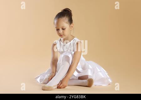 Kleine Ballerina, die spitzenschuhe vor beigefarbenem Hintergrund anzieht Stockfoto