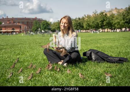 Ein junges Mädchen, das im Stadtpark auf Gras sitzt und Eisbecher hält und Spatzen füttert Stockfoto