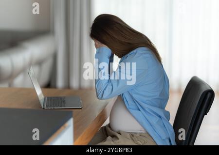Unglückliche schwangere Frau mit Kopfschmerzen, die im Heimbüro auf einem Stuhl sitzt Stockfoto