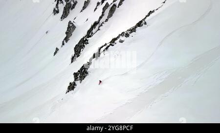Ein einzelner Snowboarder gleitet über eine steile, offene Fläche aus reinem Schnee, eingerahmt von dunklen Felsvorsprüngen. Stockfoto