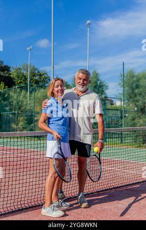 Vertikale Senioren-Tennisspieler umarmen sich glücklich, während sie ihre Schläger auf dem Clubplatz halten und direkt in die Kamera blicken Stockfoto