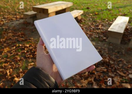 Ein weißes Buch in der Hand vor einem Hintergrund aus Holzbänken und Herbstblättern. Perfekt zur Präsentation von Bucheinbänden oder Produktverpackungen. Stockfoto