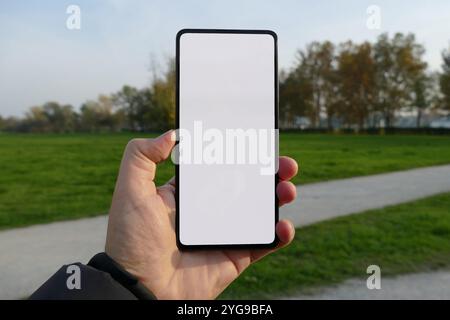 Eine Hand, die ein Smartphone mit einem leeren weißen Bildschirm vor dem Hintergrund eines Parks mit grünem Gras und Bäumen hält. Perfekt für die Präsentation mobiler App-Designs Stockfoto