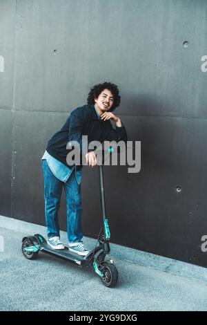 Junger Mann mit lockigen Haaren in lässigem Denim und Turnschuhen, lehnt sich auf einen schwarzen Elektroroller vor einem minimalistischen grauen Betonhintergrund. Städtisch Stockfoto