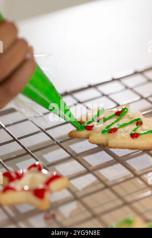 Asiatische Frau, die weihnachtskekse mit grüner und roter Glasur auf Kühlregal dekoriert, zu Hause Stockfoto